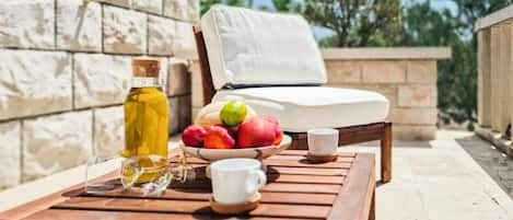 A wooden table with a bottle of olive oil, a bowl of fruit, and a white cup. A white chair with a cushion is placed behind the table.