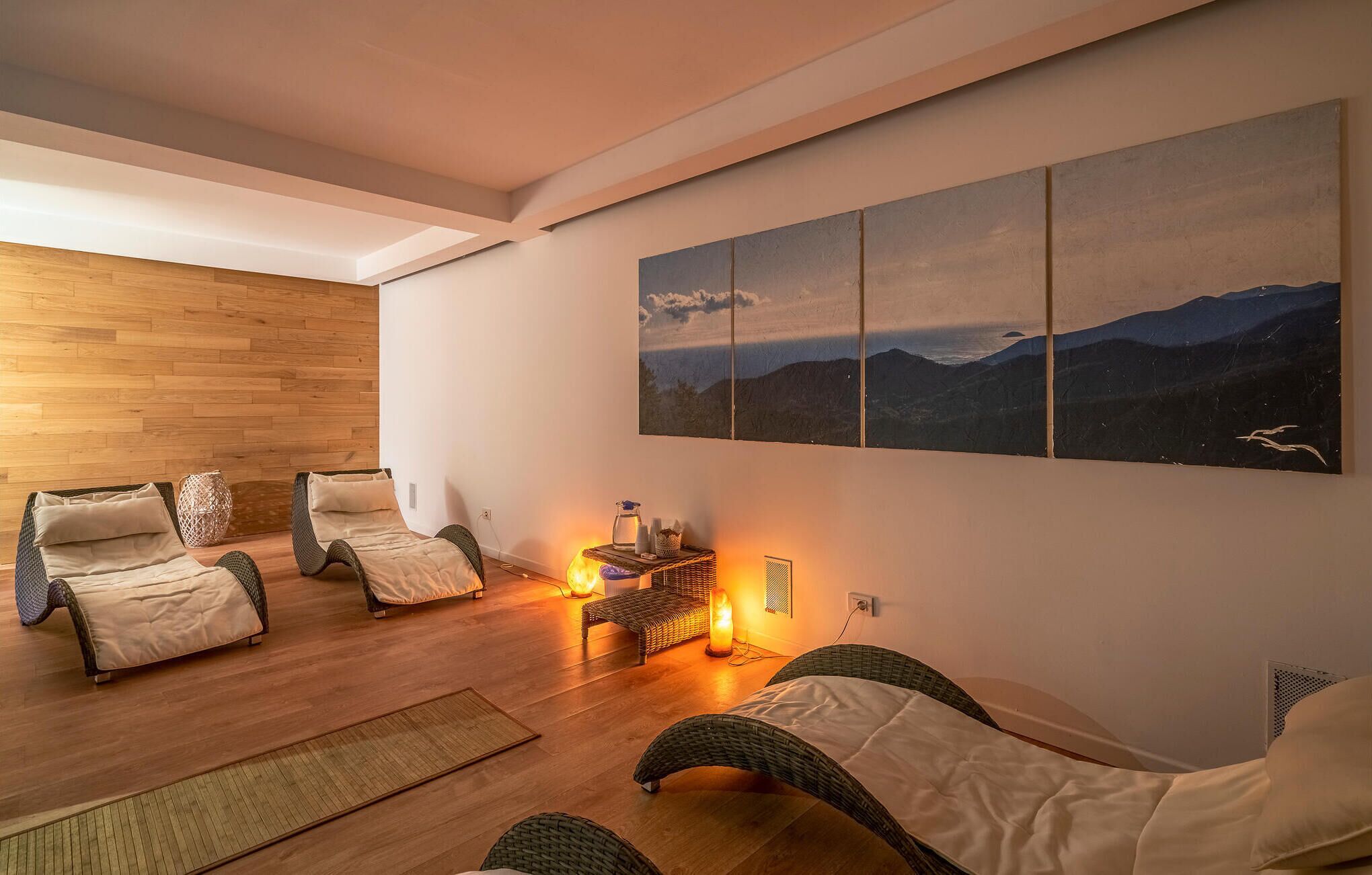 Una sala de estar moderna con piso de madera, dos sofás de mimbre, una mesita con una lámpara y un cuadro grande de paisaje en la pared.