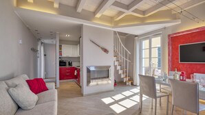 A modern living room with a sofa, dining area with chairs, a kitchen with red cabinets, a fireplace, and a staircase.