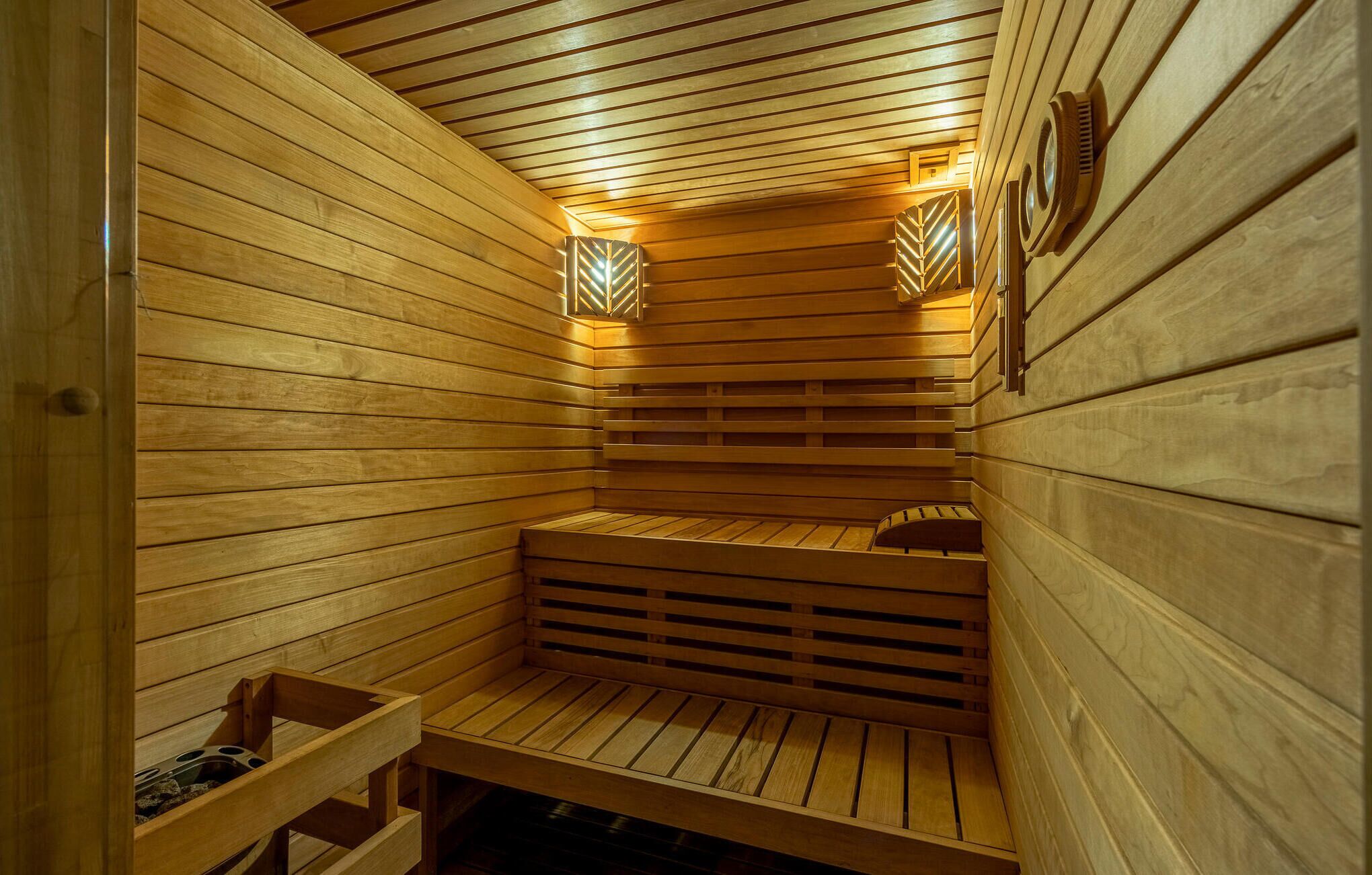 A sauna with wooden benches and slatted seating, a bench with a storage compartment, and wall-mounted lights.
