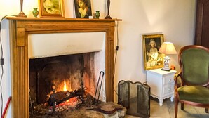 Una chimenea encendida, un sillón verde, una mesita auxiliar blanca con una lámpara, y un retrato enmarcado en la pared.