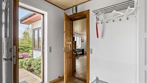 Ein Flur mit einer Holztür, die zu einem Zimmer führt, einem Kleiderständer mit Kleiderbügeln und Blick auf einen Garten durch eine geöffnete Tür.