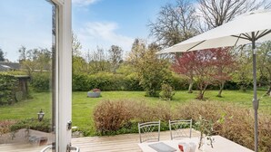 Eine Terrassengruppe im Freien mit Tisch, Stühlen und einem weißen Sonnenschirm, mit Blick auf einen Garten mit Bäumen und Rasen.