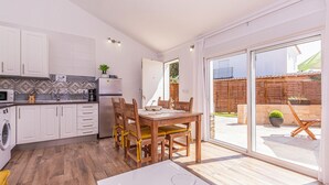 Una cocina moderna con armarios blancos, microondas y cafetera. Hay un comedor con mesa y sillas de madera, y un amplio ventanal corredera que da a un patio con cerca de madera y plantas.