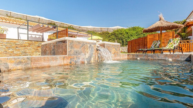 Una piscina cristalina con caída de agua, rodeada de muros de piedra y muebles de madera.