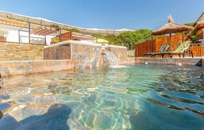 Una piscina cristalina con caída de agua, rodeada de muros de piedra y muebles de madera.