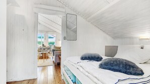 Et soveværelse med trægulv, seng med hvidt sengetøj og blå detaljer, et spejl på væggen, samt udsigt til et udendørs område med et bord og stole.