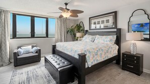 A bedroom with a large bed, a ceiling fan, a nightstand, a bench, and a view of the ocean through large windows. - 2/2 OCEANFRONT 8th Floor Unit with Open Pool (Daytona Beach)