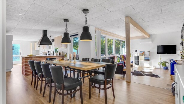 A spacious dining area with a long wooden table and multiple chairs, a kitchen area with white cabinets, and a large window providing natural light.