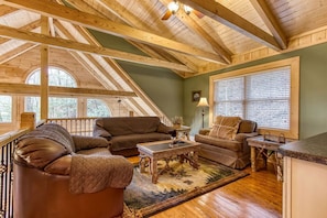 A cozy living room with a wooden ceiling, a large window with blinds, a brown leather sofa, a wooden coffee table, and a small side table. - Running Bears Den- Chalet Village’s Perfect Retreat in the Mountains (Gatlinburg)