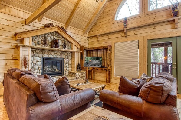 A cozy log cabin interior with a stone fireplace, leather sofas, and a television. - Running Bears Den- Chalet Village’s Perfect Retreat in the Mountains (Gatlinburg)