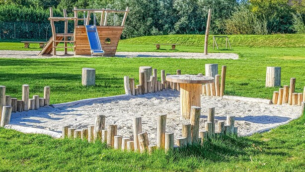 Une aire de jeux avec un toboggan en bois et des structures d’escalade, entourée de sable et d’herbe.