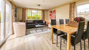 Un salon moderne avec un canapé, une table basse et une table à manger avec des chaises. Il y a une grande fenêtre avec des rideaux, un tableau au mur et une vue sur la verdure à l’extérieur.