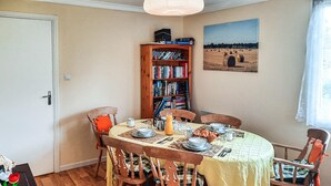 Un coin repas avec une table ronde dressée pour quatre personnes, entourée de chaises en bois. Une bibliothèque remplie de livres est adossée au mur, et un tableau représentant un paysage est accroché au-dessus de la table.
