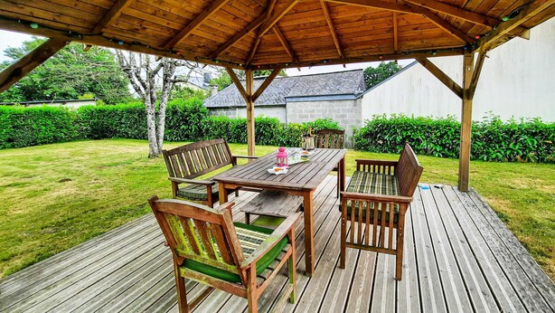 Une terrasse en bois avec une table et des chaises, entourée de verdure et avec une maison en arrière-plan.