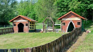 Dos estructuras de madera rústica con entradas en arco, rodeadas por una valla de madera, ubicadas en un área cubierta de césped con árboles al fondo.