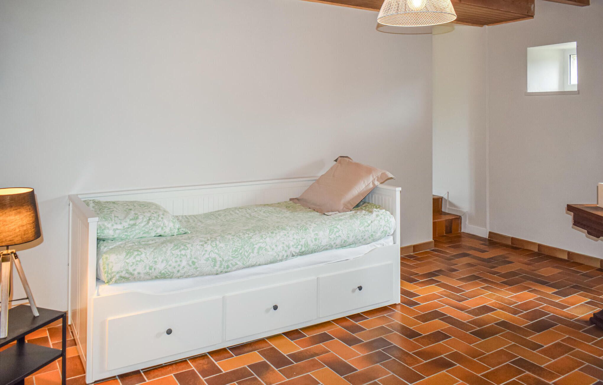 A bedroom with a white bed, a lamp, a chest of drawers, and a herringbone-patterned floor.