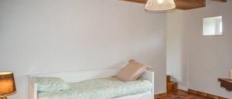A bedroom with a white bed, a lamp, a chest of drawers, and a herringbone-patterned floor.