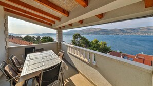 Varanda com mesa e cadeiras, vista para um corpo de água com barcos e um horizonte urbano ao longe.