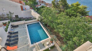 Uma piscina no terraço com vista para o mar, rodeada por vegetação e edifícios.