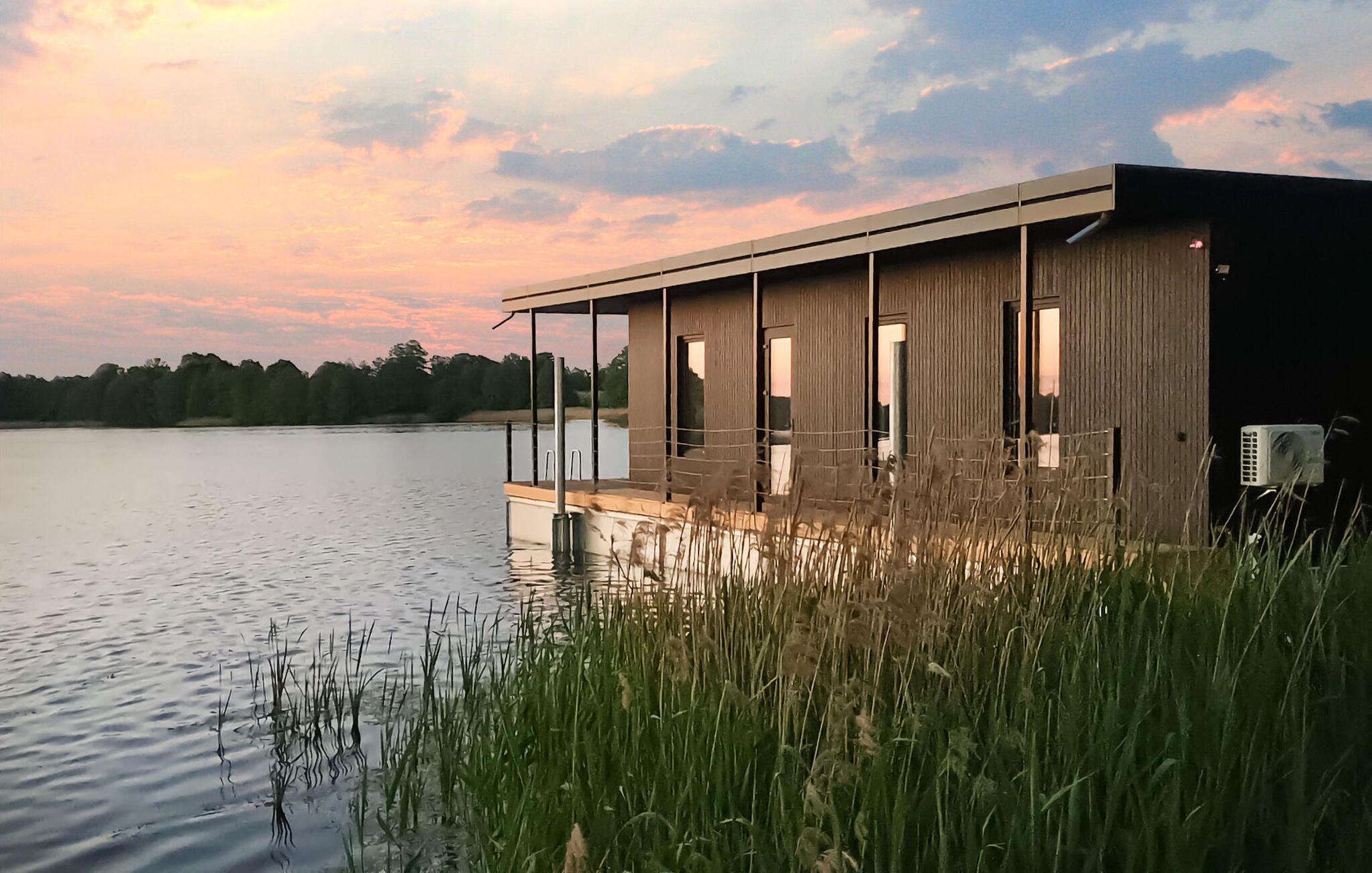 Un bâtiment moderne de style cabane, doté d’une terrasse qui s’étend au-dessus d’une étendue d’eau, et entouré de hautes herbes.