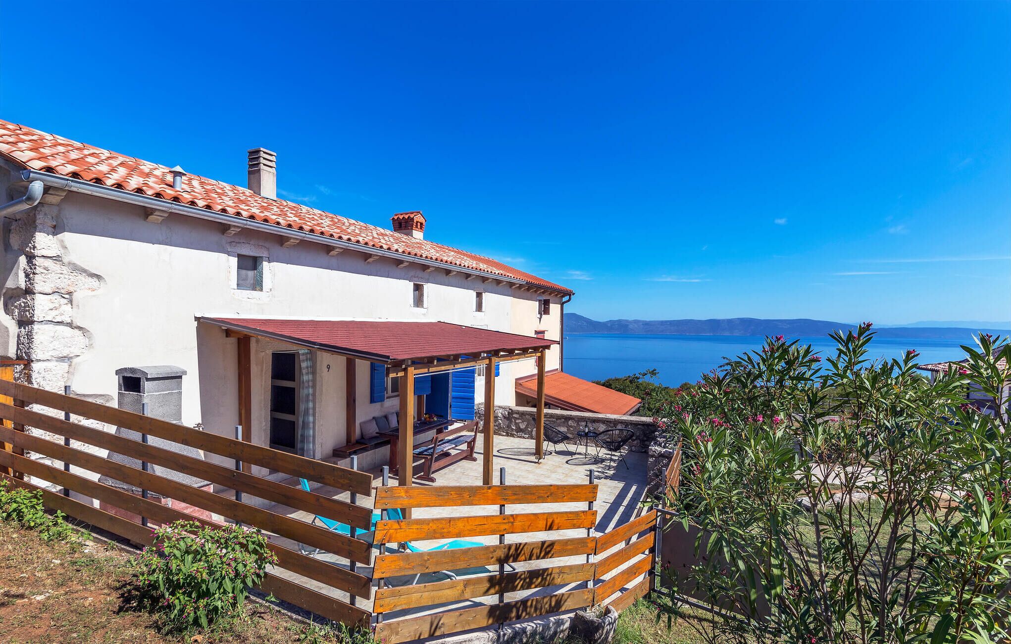A white building with a red-tiled roof and a wooden fence. It has a view of the sea and mountains in the background.
