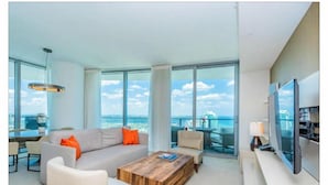A modern living room with a large sofa, coffee table, and a TV. The room has large windows offering a view of the city and the sea. - Hyde Resort Hollywood Full Amenities BOOK NOW! (Hollywood)