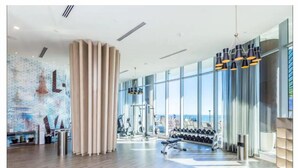 A modern gym with exercise equipment, large windows, and a brick wall mural. - Hyde Resort Hollywood Full Amenities BOOK NOW! (Hollywood)