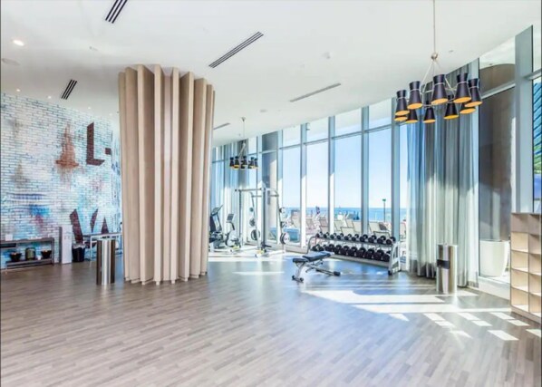 A modern gym with exercise equipment, large windows, and a brick wall mural.