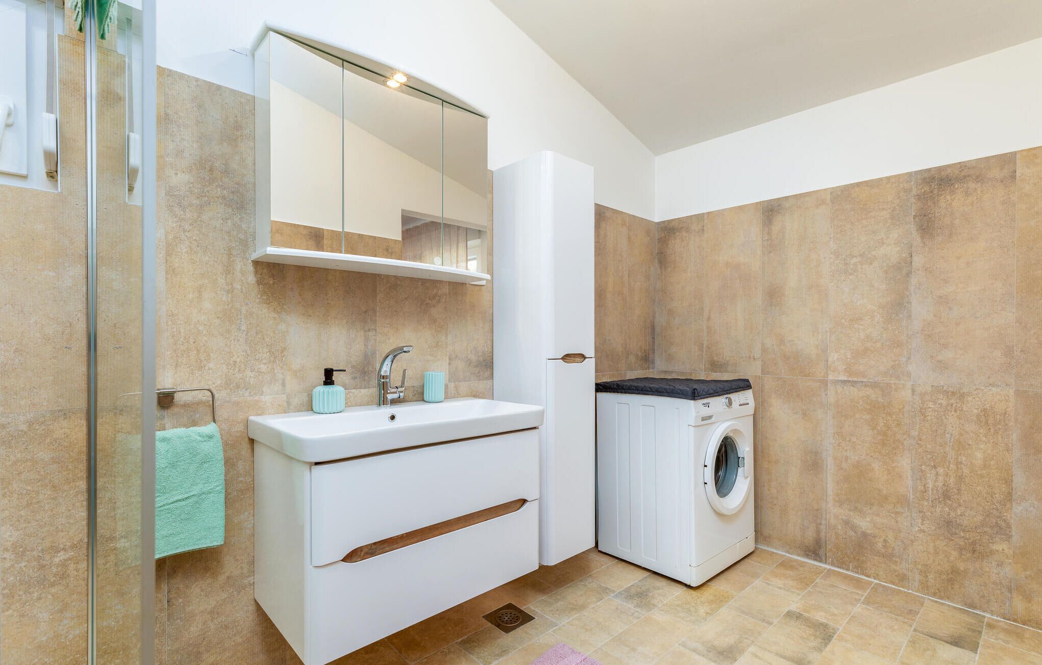 A modern bathroom with a glass shower enclosure, a white sink with a mirror above, and a front-loading washing machine.