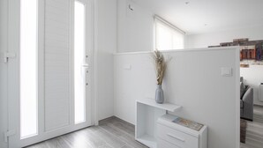 Ett modernt, minimalistiskt rum med en skjutdörr i glas som leder ut till en balkong, en vit hylla med en vas och en grå soffa i bakgrunden.