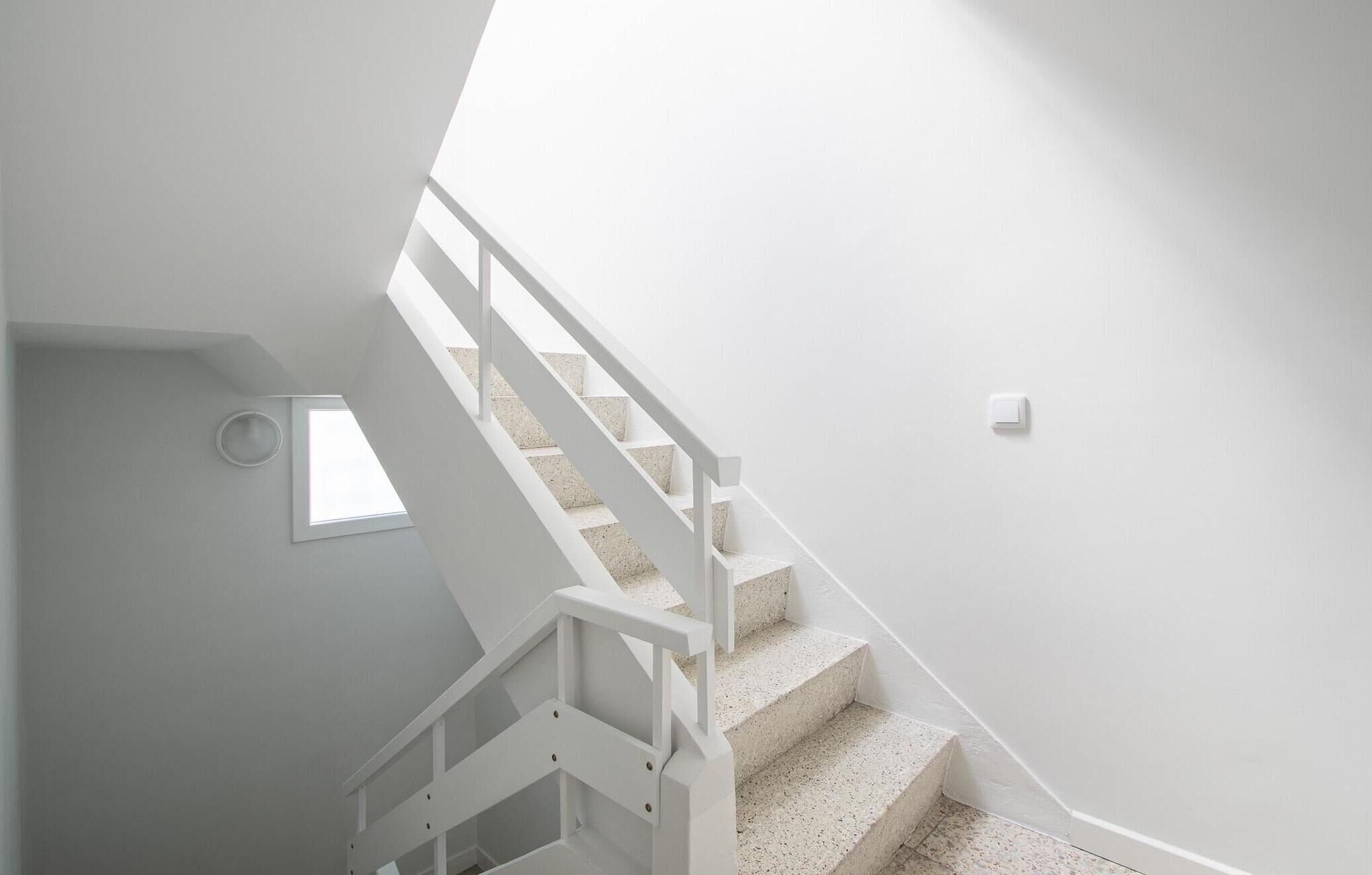 Un escalier blanc avec une rampe, menant à un palier doté d’une petite fenêtre.