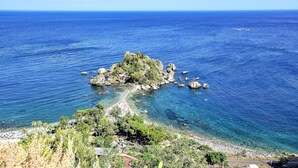 Uma área costeira com uma ilha rochosa, mar azul cristalino e uma praia com guarda-sóis e árvores.