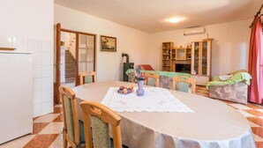 Uma sala de jantar com uma mesa posta para quatro pessoas, rodeada por cadeiras de madeira. Um refrigerador é visível à esquerda, e uma escada leva a um andar superior.