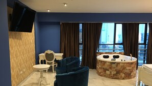 Une chambre d’hôtel moderne avec une baignoire en pierre, un fauteuil bleu, un bureau blanc et un téléviseur fixé au mur.
