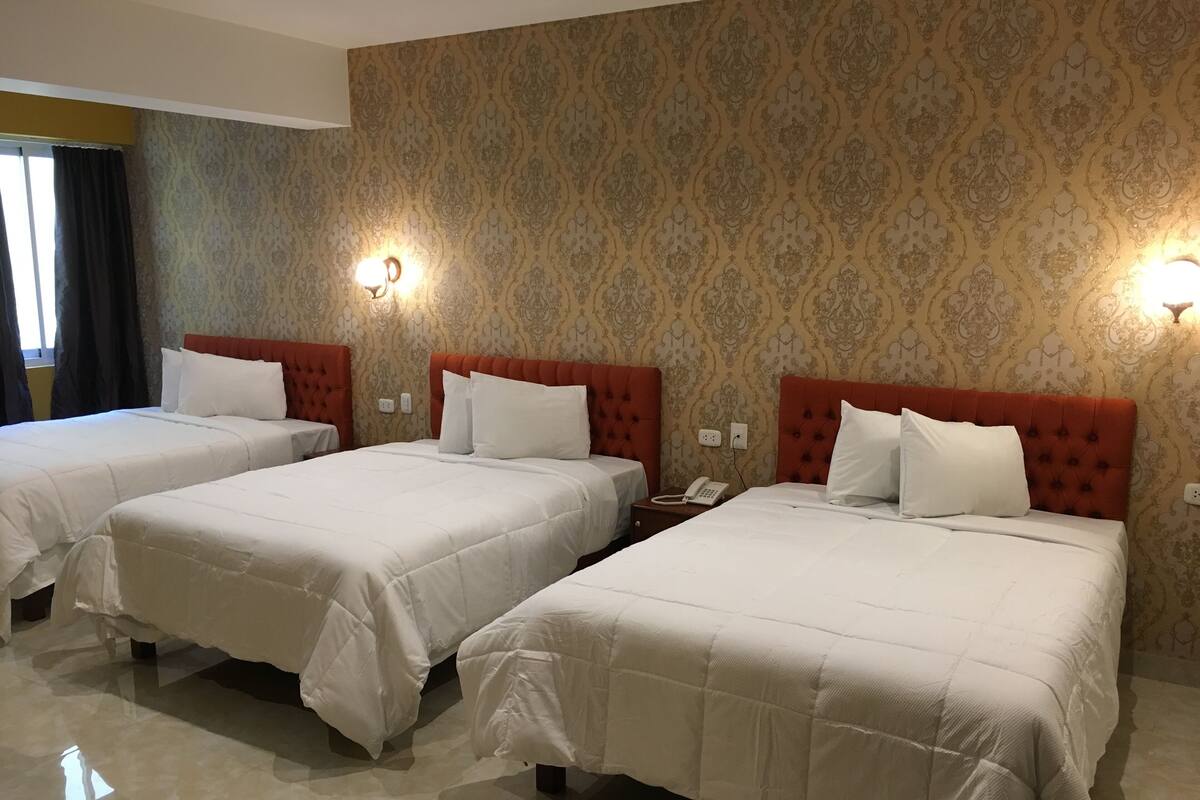 A hotel room with three beds, each with white bedding and red headboards. There are wall-mounted lights and a telephone on the nightstand.