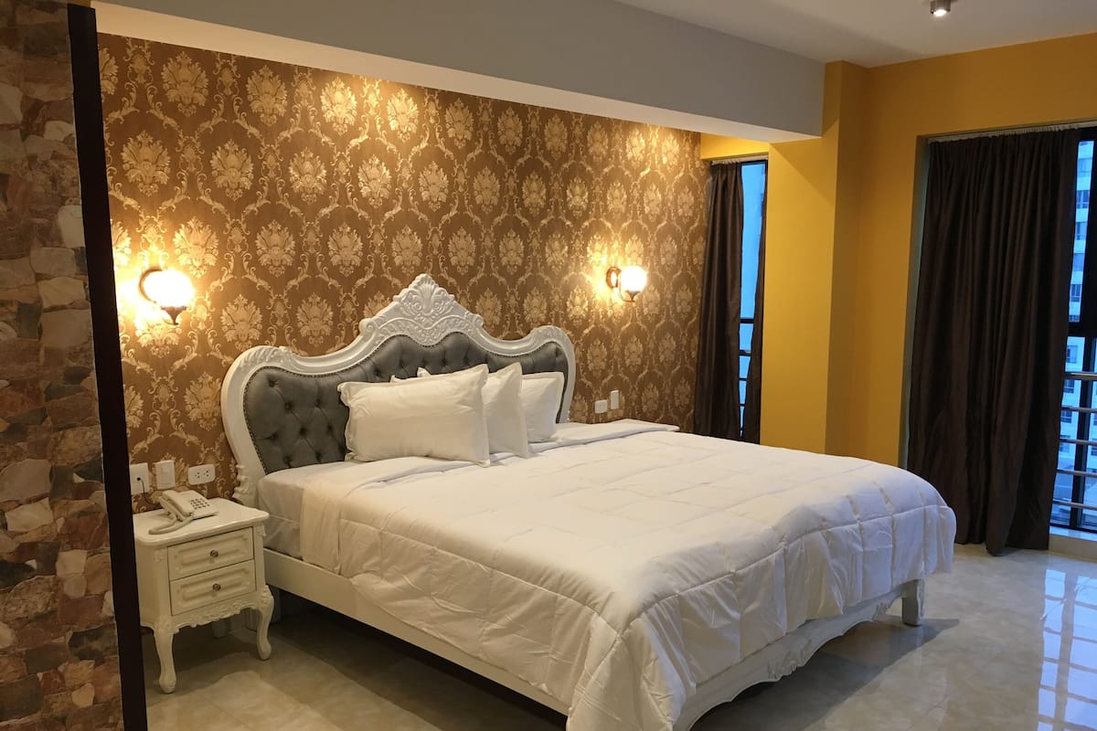 A neatly made bed with a headboard, a bedside table with a phone, and a wall with patterned wallpaper.