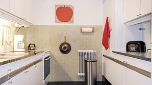 A kitchen with white cabinets, a coffee machine, a kettle, a frying pan, a radiator, and a trash bin. - Stunning apartment in Bergen aan Zee (Bergen aan Zee)