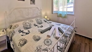 A bedroom with a bed featuring a nautical-themed bedspread, a white headboard, a bedside table with a lamp, and a window with a sheer curtain. - Amazing home in Risika (Risika)