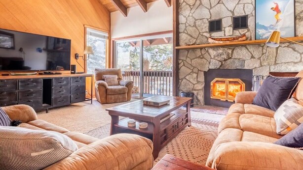 Un salon chaleureux avec une cheminée en pierre, des poutres apparentes en bois au plafond, un téléviseur et des fauteuils confortables.