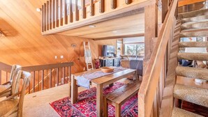 Un intérieur en bois avec un plafond haut, un escalier en bois et un coin repas agrémenté d’un tapis à motifs rouges.