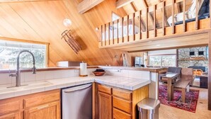 Une cuisine moderne avec des armoires en bois, un lave-vaisselle et une cuisinière encastrée. On y trouve un coin repas avec une table et des chaises, et une vue sur l’extérieur grâce à de grandes fenêtres.