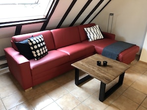 Un canapé rouge agrémenté de deux coussins à motifs, une table basse en bois sur laquelle reposent deux tasses noires, et un lampadaire.