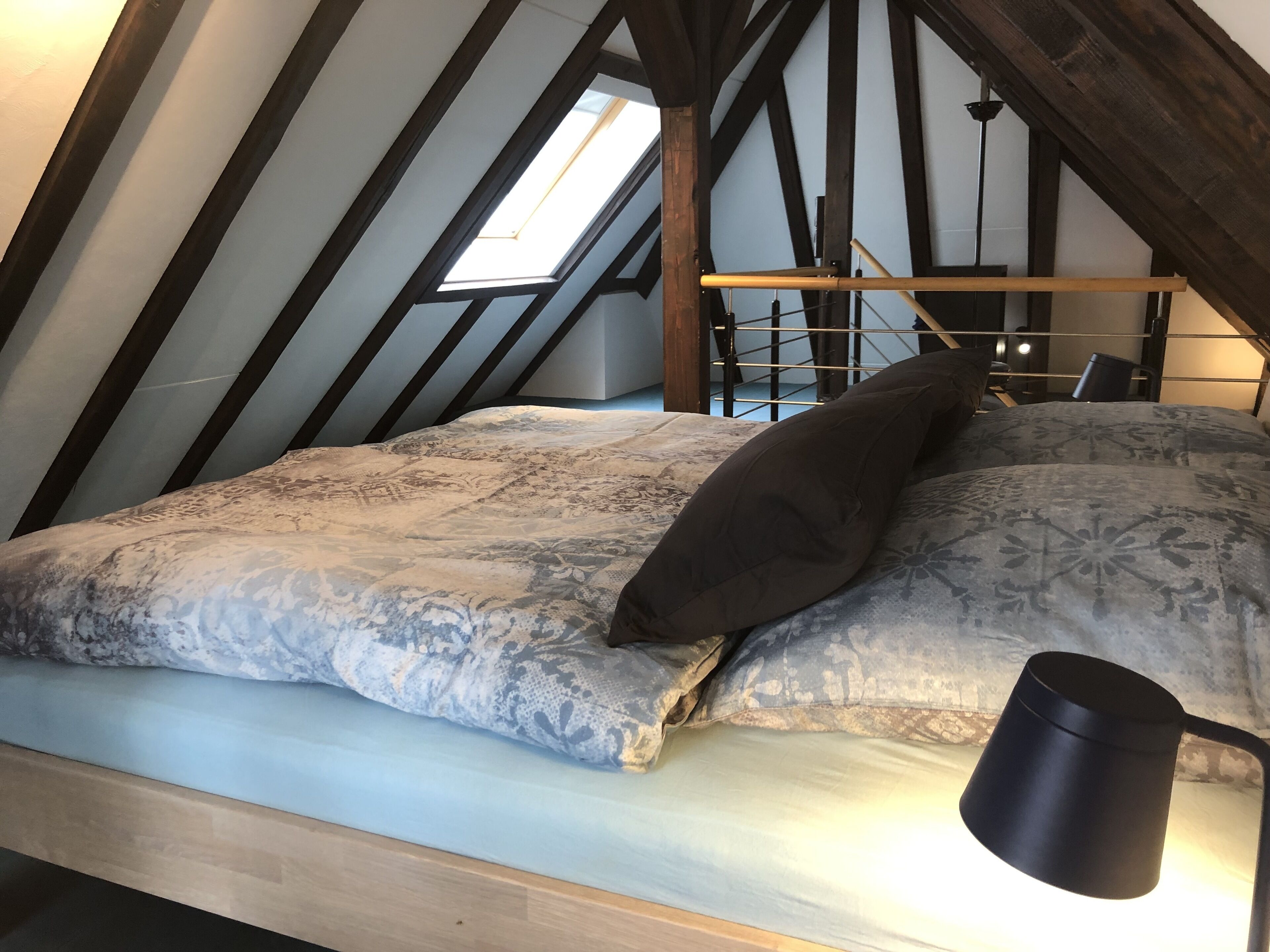 Une chambre de style loft avec un plafond en pente, un puits de lumière, un lit avec une literie à motifs, une lampe de chevet et une tête de lit en bois.