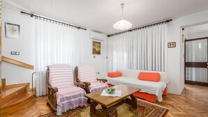 A living room with a wooden staircase, a sofa with orange cushions, striped chairs, a wooden coffee table, and a chandelier. - Beach front home in Barbat na Rabu (Barbat na Rabu)
