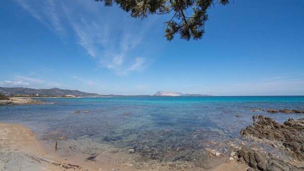 Een strand met helder turquoise water, een rotsachtige kustlijn en bergen in de verte.
