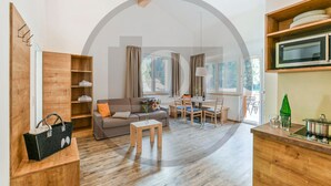 A modern living room with a sofa, coffee table, and dining area. There is a wooden bookshelf, a kitchen area with a microwave, and a large window providing natural light. - schoene Ferienwohnung (Klösterle)