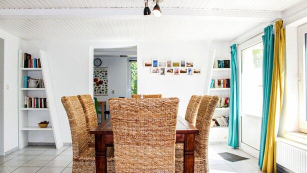 Ein Essbereich mit Korbstühlen und einem Holztisch, umgeben von Bücherregalen und Blick ins Freie.