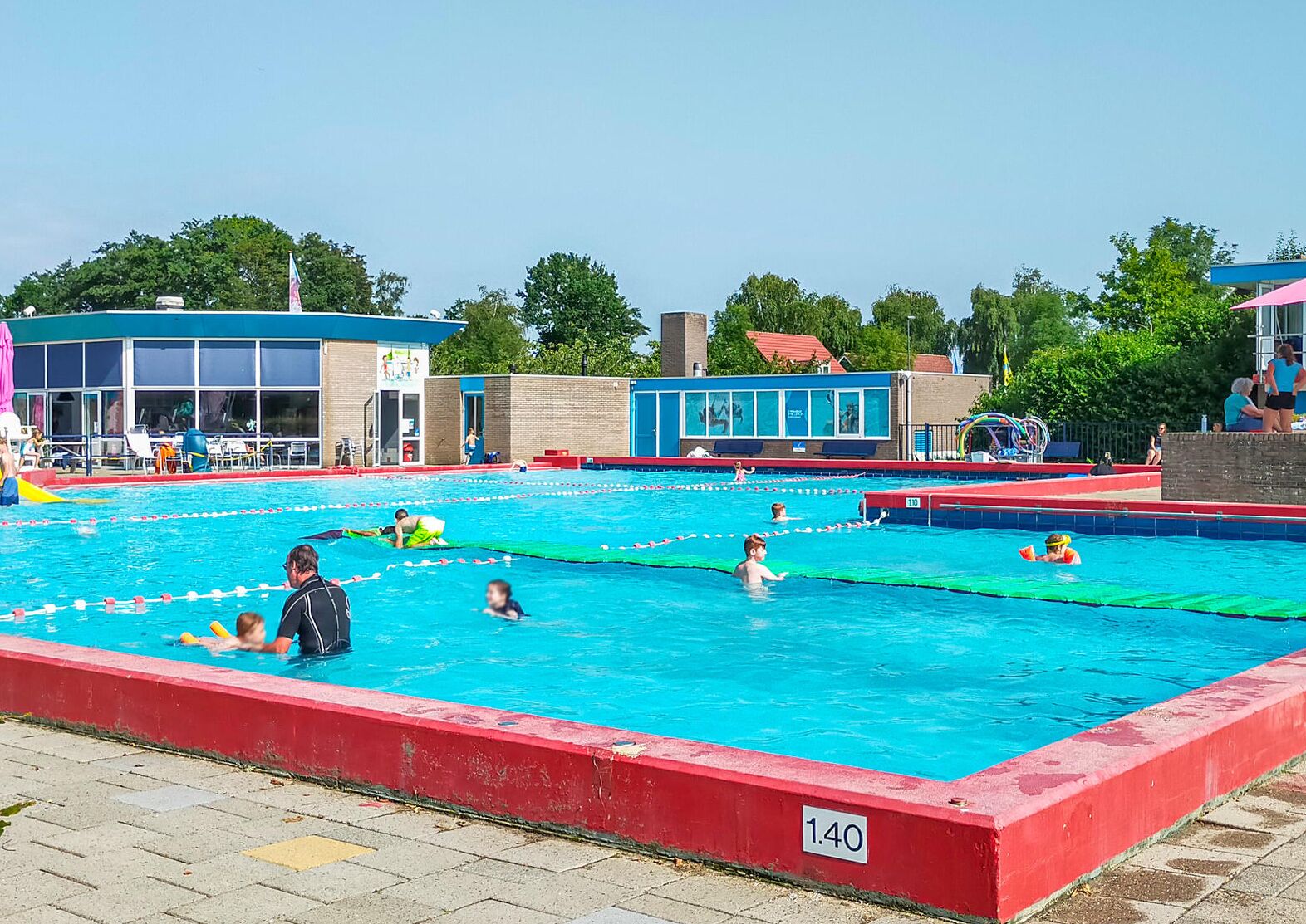 A public swimming pool with multiple lanes, a lifeguard tower, and a building with a blue roof in the background.