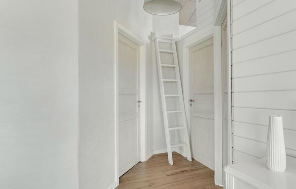 Un couloir étroit avec un plancher en bois et des murs blancs. Une échelle blanche est appuyée contre l’un des murs, et un vase blanc est posé sur une table dans un coin.